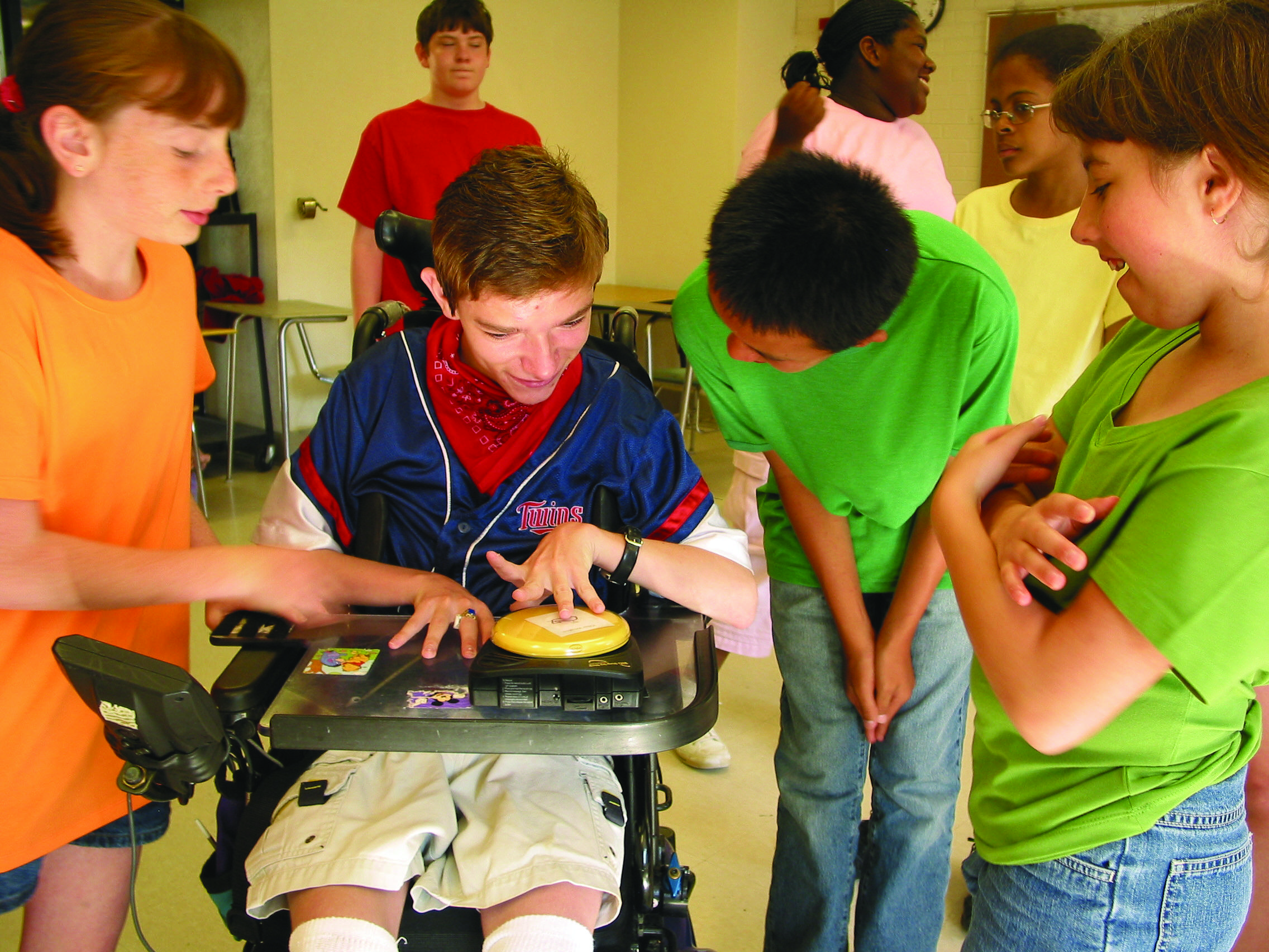 Image of several kids talking. One of the kids is seated on a powered wheelchair and using a single switch. 
